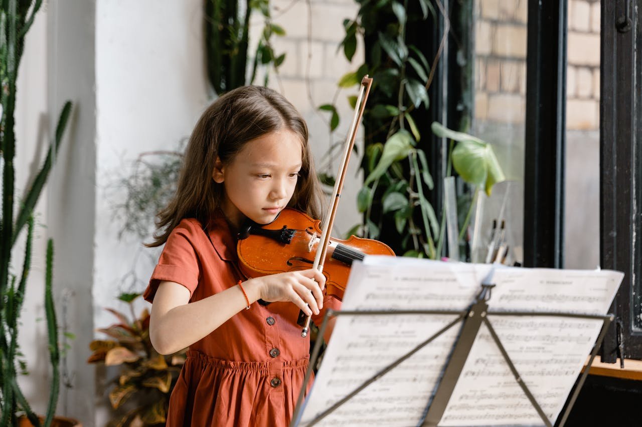A young girl plays the violin with sheet music, focusing intently in an indoor setting.